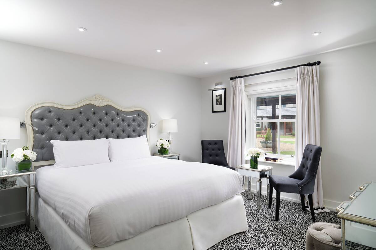 A hotel room with a large bed, a bedside table with a lamp, a chair, a small table with a vase of flowers, and a window with curtains.