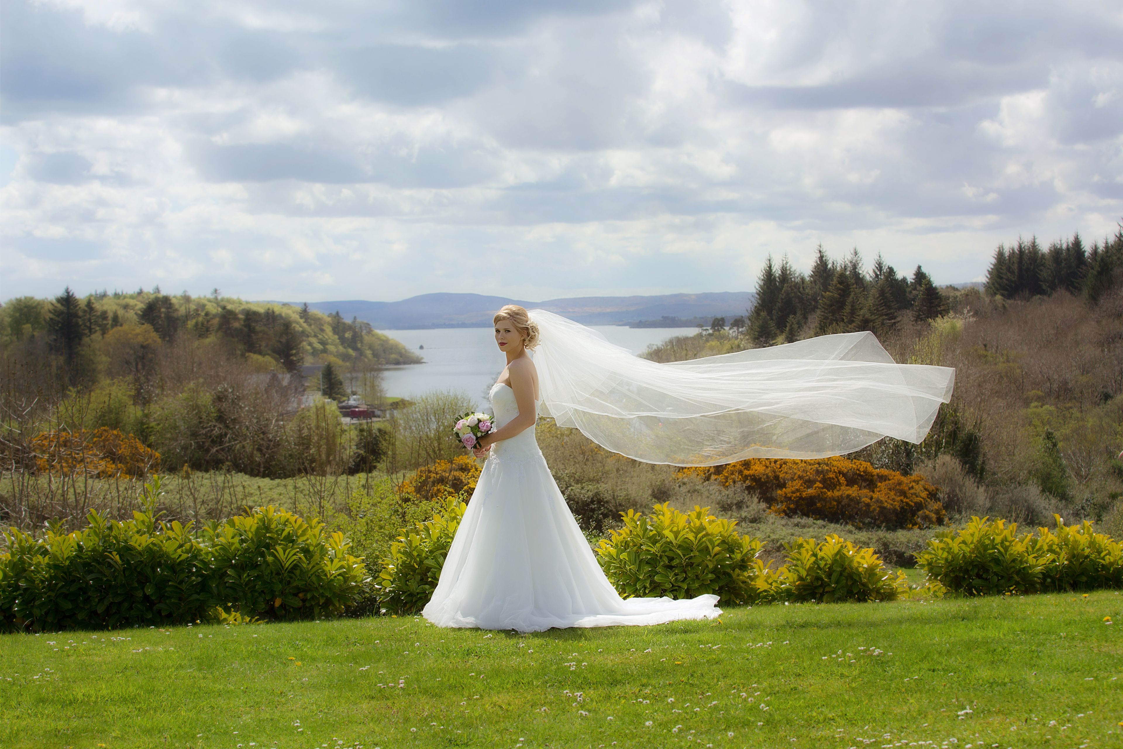 A bride in a white wedding dress with a long veil, standing on a grassy field with a scenic view of a lake and mountains in the background.