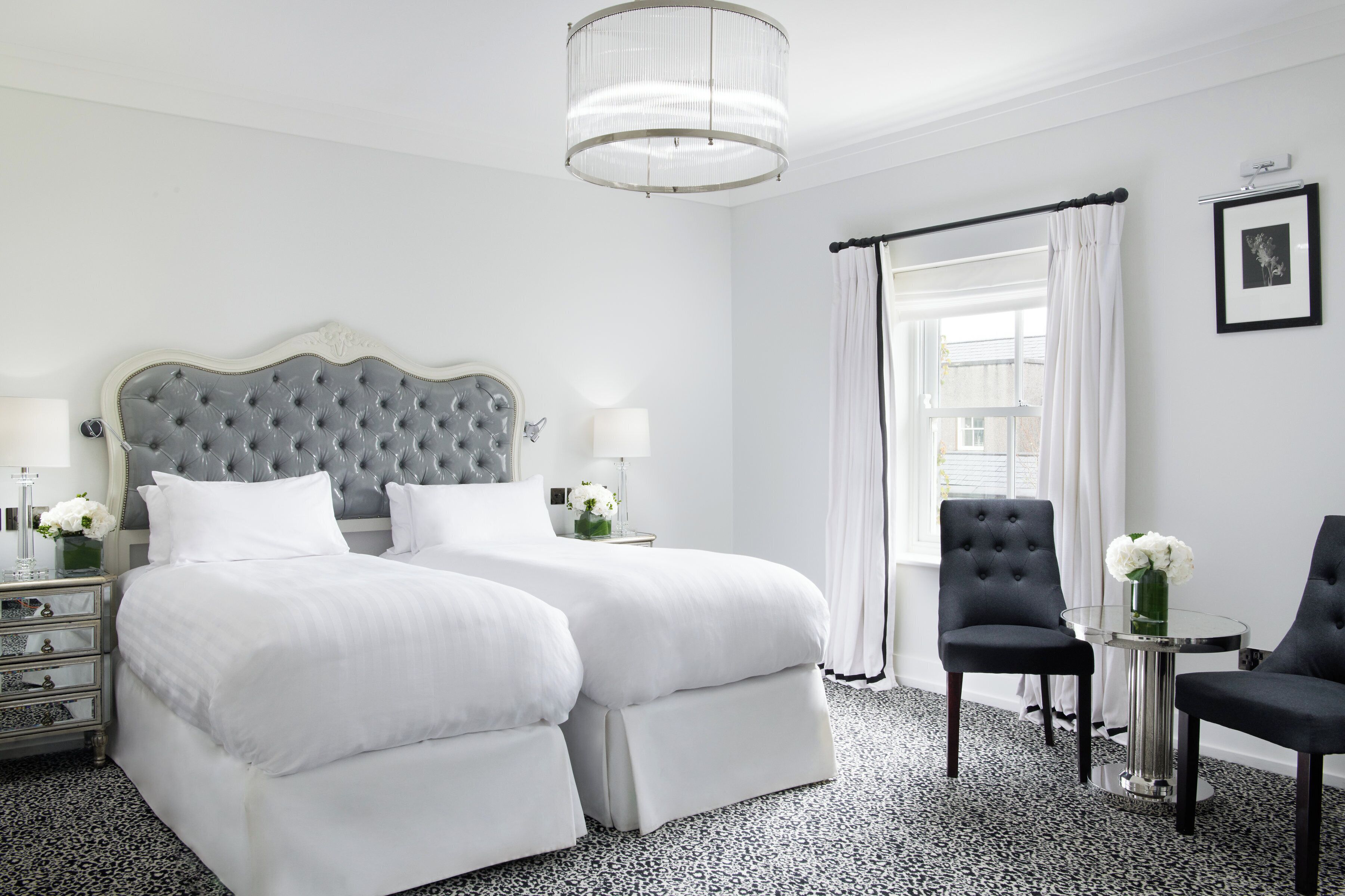 A hotel room with a large bed, two bedside tables, a chair, a small table with a vase, and a chandelier.