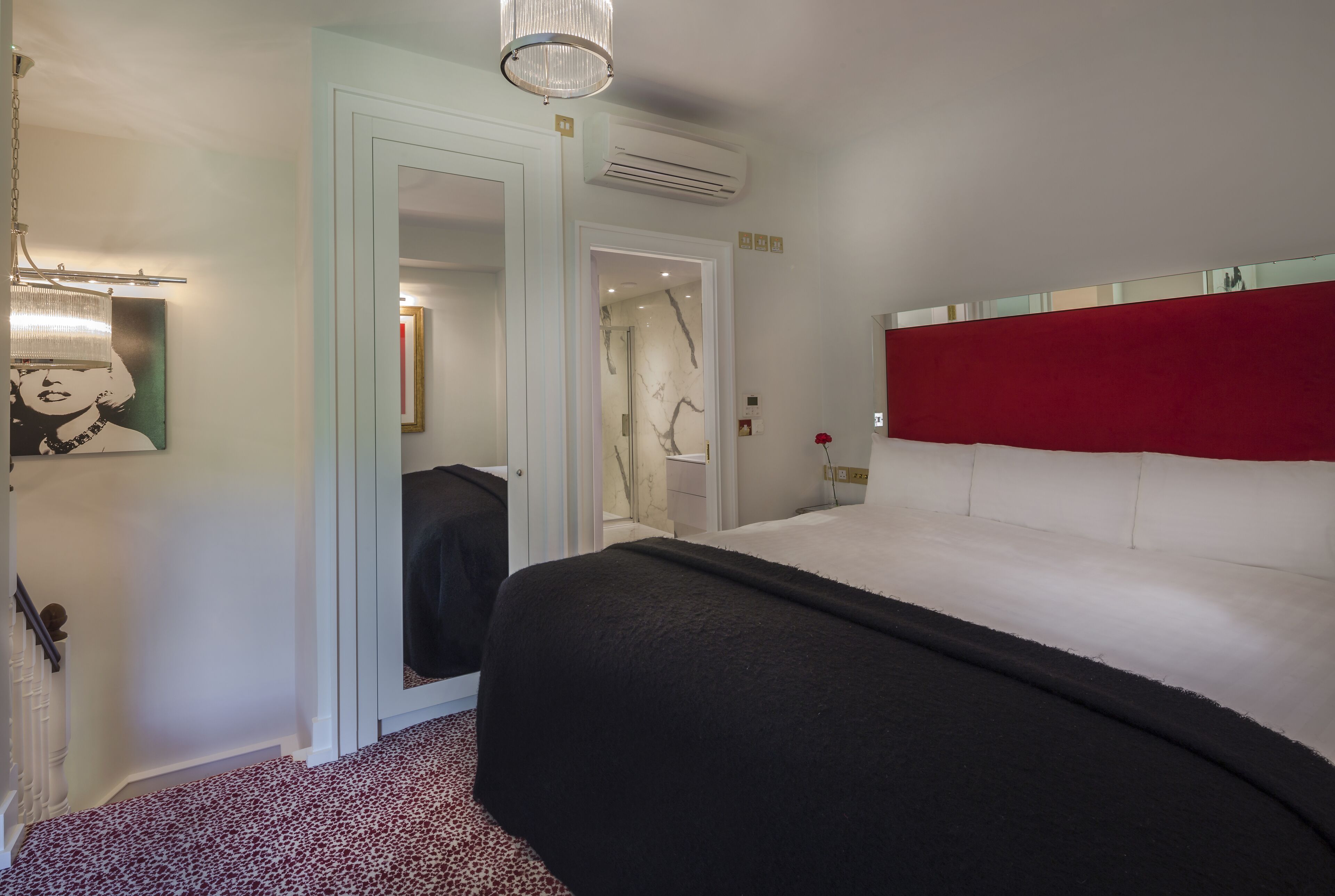 Une chambre à coucher avec un lit, un miroir, une porte donnant accès à une salle de bain et une tête de lit rouge.