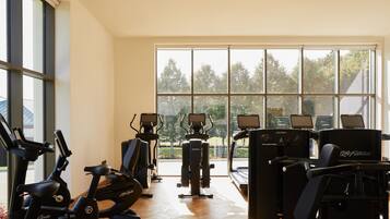 Un gimnasio con máquinas de cardio, equipo de entrenamiento de pesas y amplios ventanales con vistas a árboles.