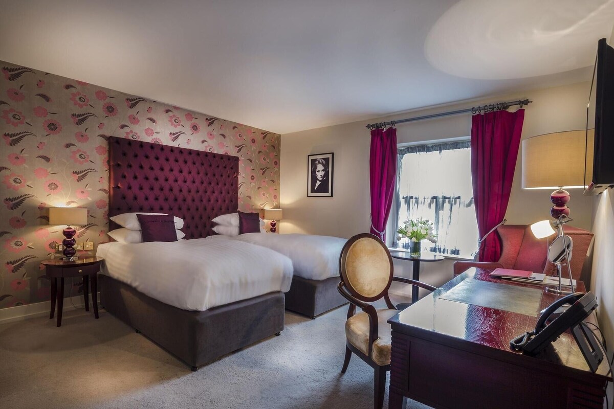 Ein Hotelzimmer mit zwei Betten, einem Schreibtisch, einem Stuhl, einem Fernseher und einem Fenster mit Vorhängen.