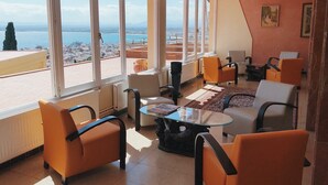 Amplio salón con grandes ventanales que ofrecen vistas a la ciudad, mobiliario moderno color naranja y una mesa de centro de vidrio.
