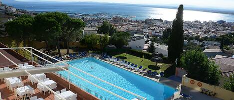 A rooftop pool with lounge chairs and tables, overlooking a coastal cityscape.