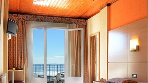 Habitación de hotel con techo de madera, cama con ropa de cama floral, televisor, silla, escritorio y balcón con vista al mar.