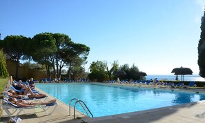 A pool area with lounge chairs and umbrellas, surrounded by trees and a view of the sea. - Hotel Grecs (Roses)