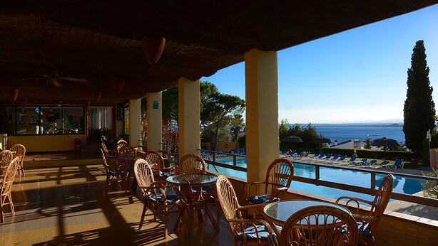 Un espacio exterior con asientos de mimbre y mesas, con vista a una piscina y al mar.