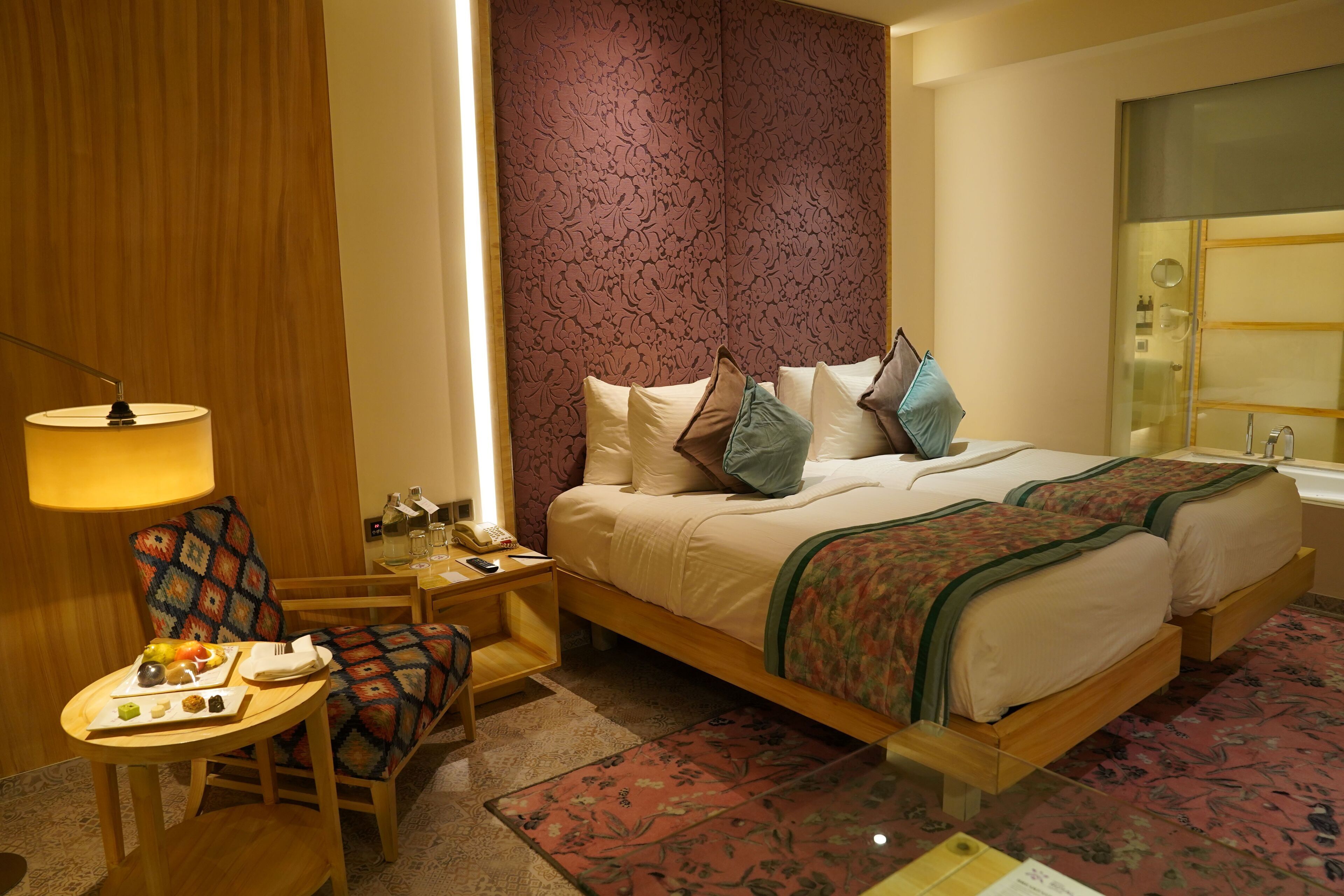 Ein Hotelzimmer mit Doppelbett, Nachttisch mit Lampe, Sessel und kleinem Tisch mit Snacks.