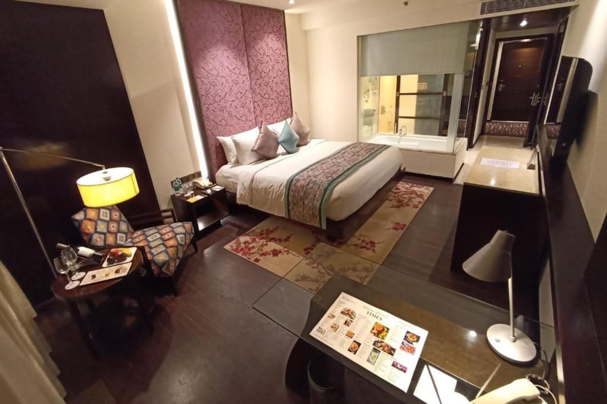 Quarto de hotel com cama, mesa de cabeceira, abajur, cadeira e área de refeições com menu.