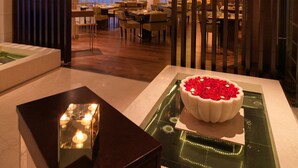 Un vestíbulo de hotel con una recepción, un comedor con mesas y sillas, y un centro de mesa decorativo con flores rojas.