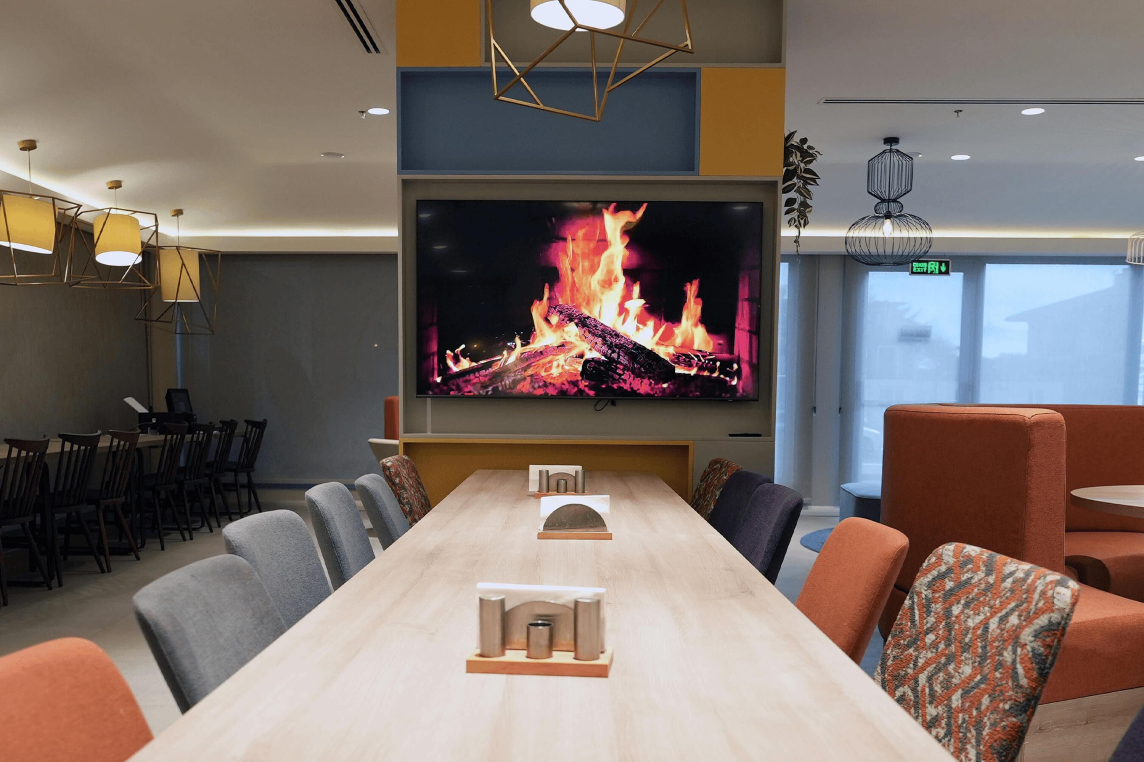 A conference room with a long table, chairs, a TV screen displaying a fire, and a modern ceiling design.
