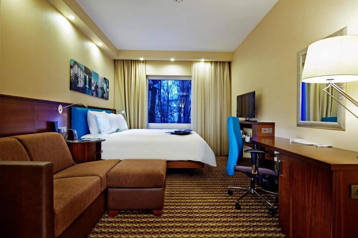 Habitación de hotel con cama, escritorio, sillón, sofá y ventana grande con vista a árboles.