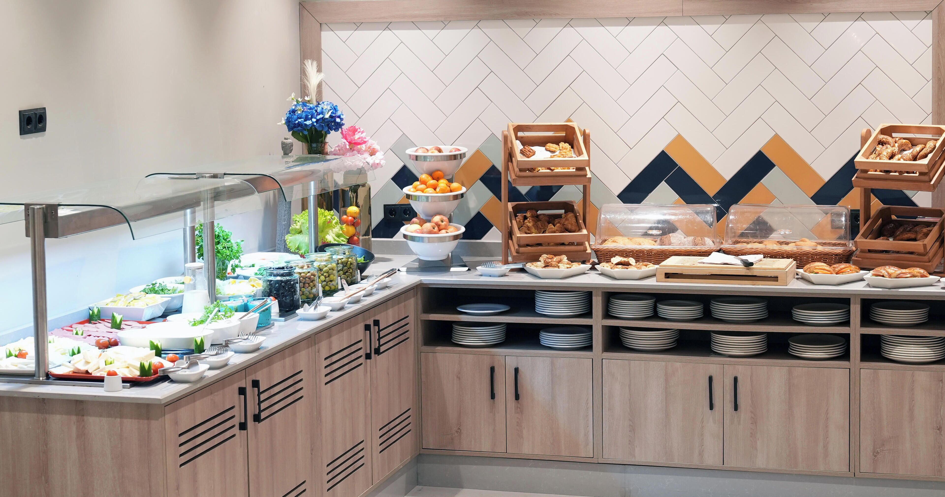 Una mesa de buffet con diversas opciones de comida, como frutas, pasteles y ensaladas, dispuestas sobre un mostrador con armarios de madera y salpicadero de azulejos.