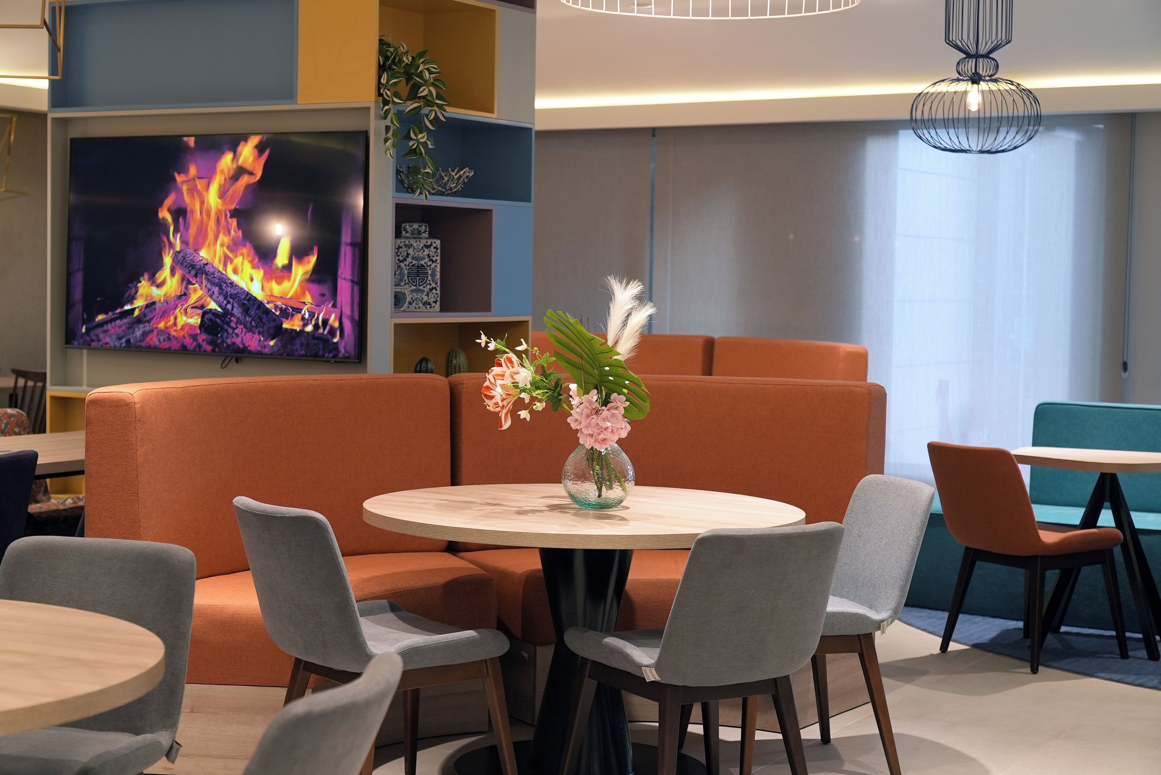 Un área de estar moderna con asientos naranja, una mesa redonda con sillas, un televisor de pantalla plana mostrando un fuego y un jarrón decorativo con flores.