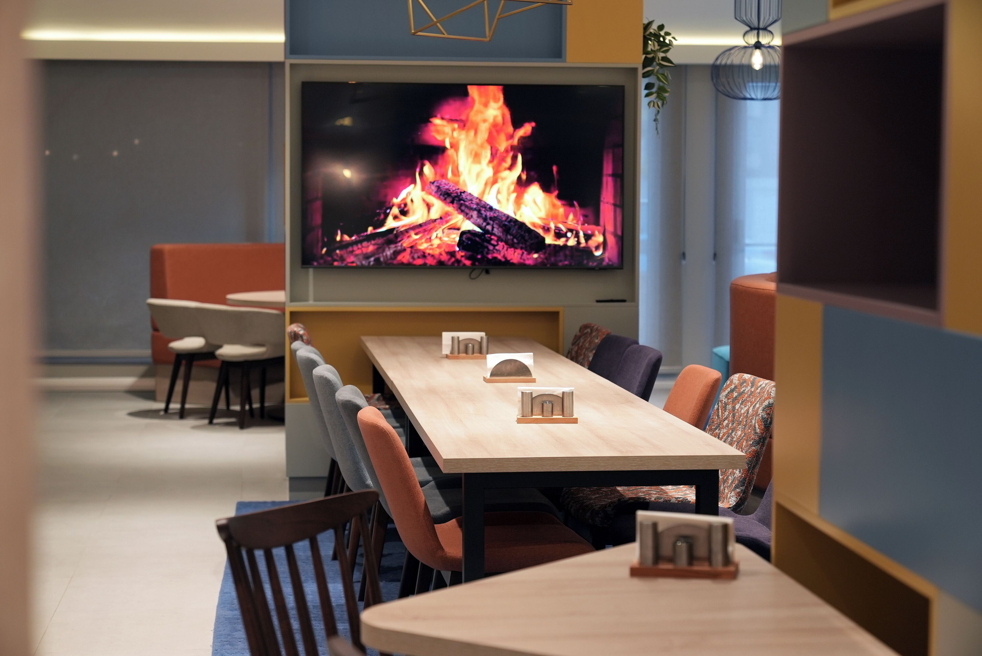 A modern meeting room with a large flat-screen TV displaying a fire, wooden tables, and comfortable seating.