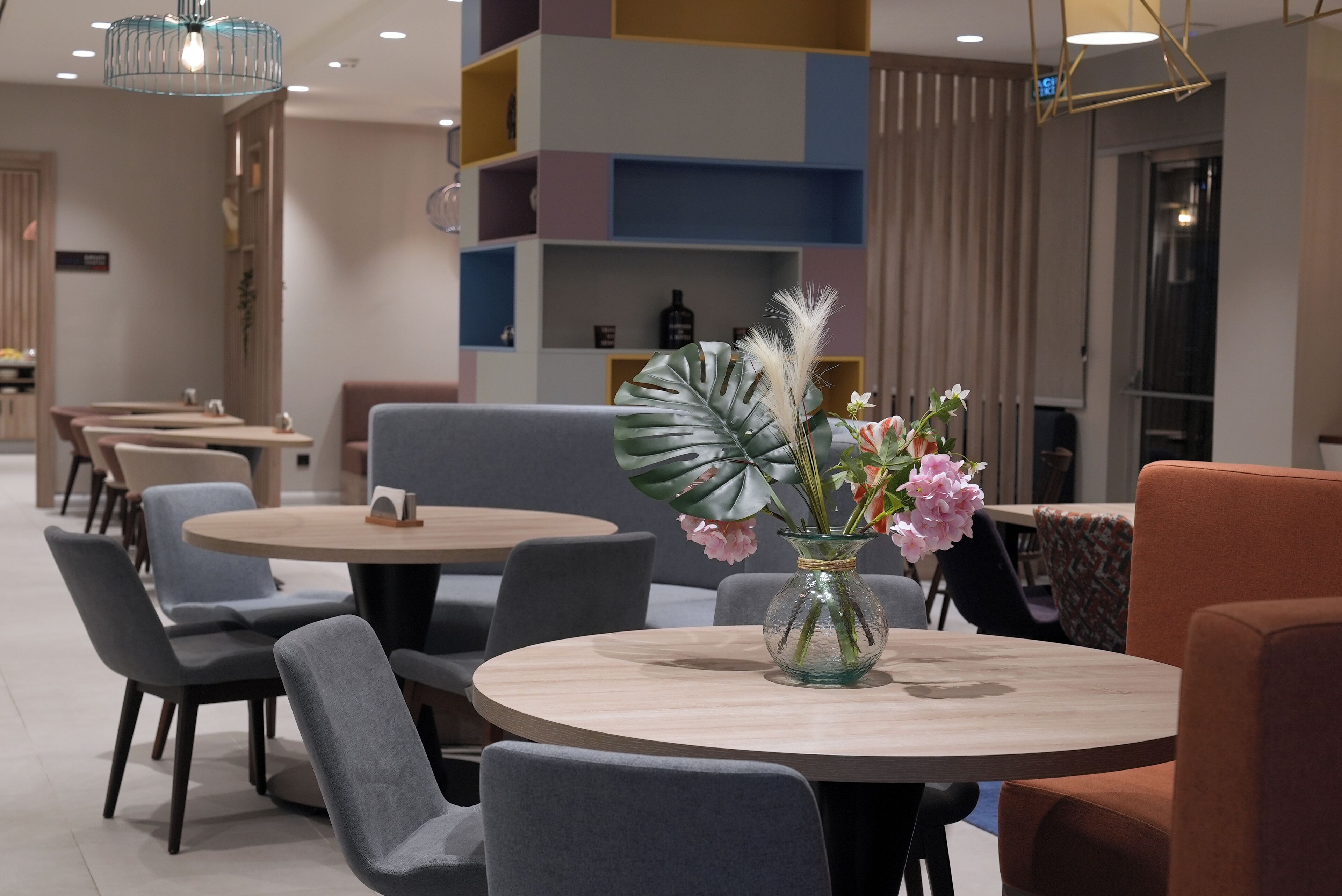 A modern dining area with round tables and grey chairs, a vase with flowers, and a colorful shelving unit in the background.