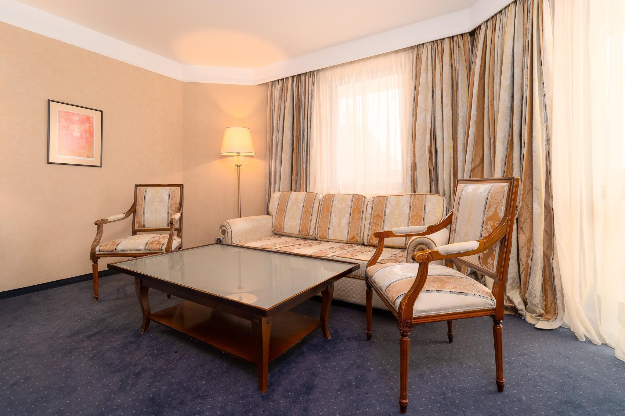 A hotel room with a sofa, two armchairs, a glass coffee table, a floor lamp, and a large window with curtains.
