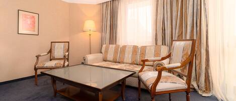 A hotel room with a sofa, two armchairs, a glass coffee table, a floor lamp, and a large window with curtains.