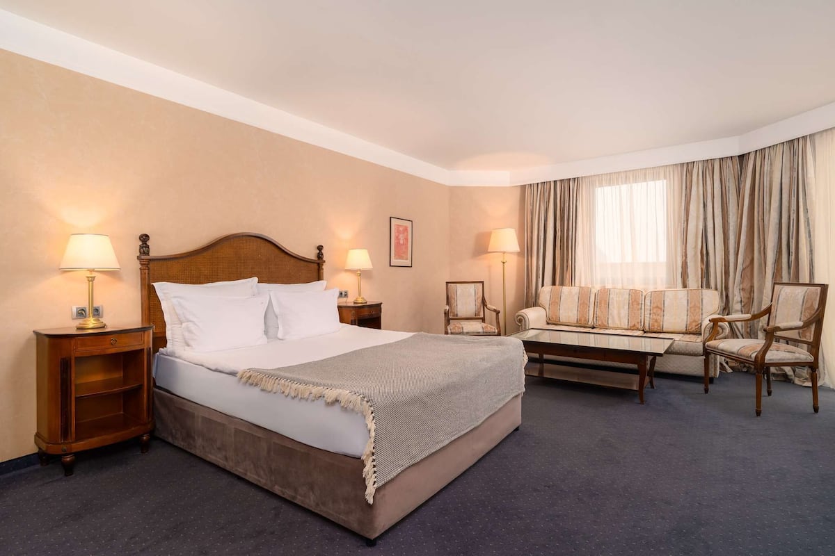 A hotel room with a large bed, two bedside tables with lamps, a sofa, two armchairs, and a window with curtains.