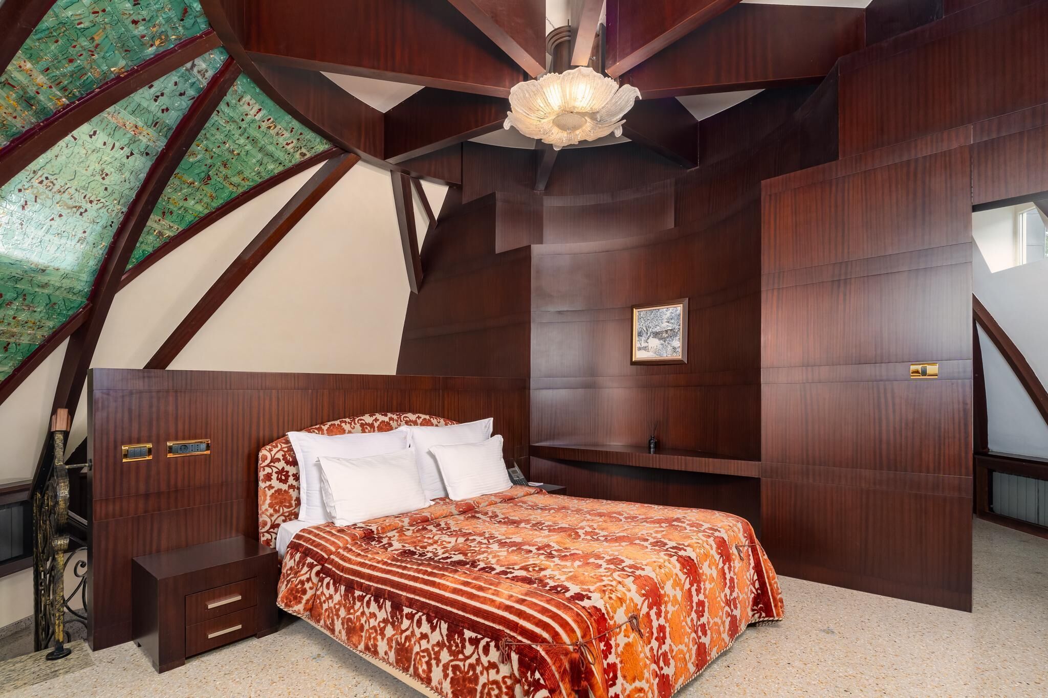 Un dormitorio con cabecera de madera, cama con colcha estampada, mesita de noche y lámpara de techo.