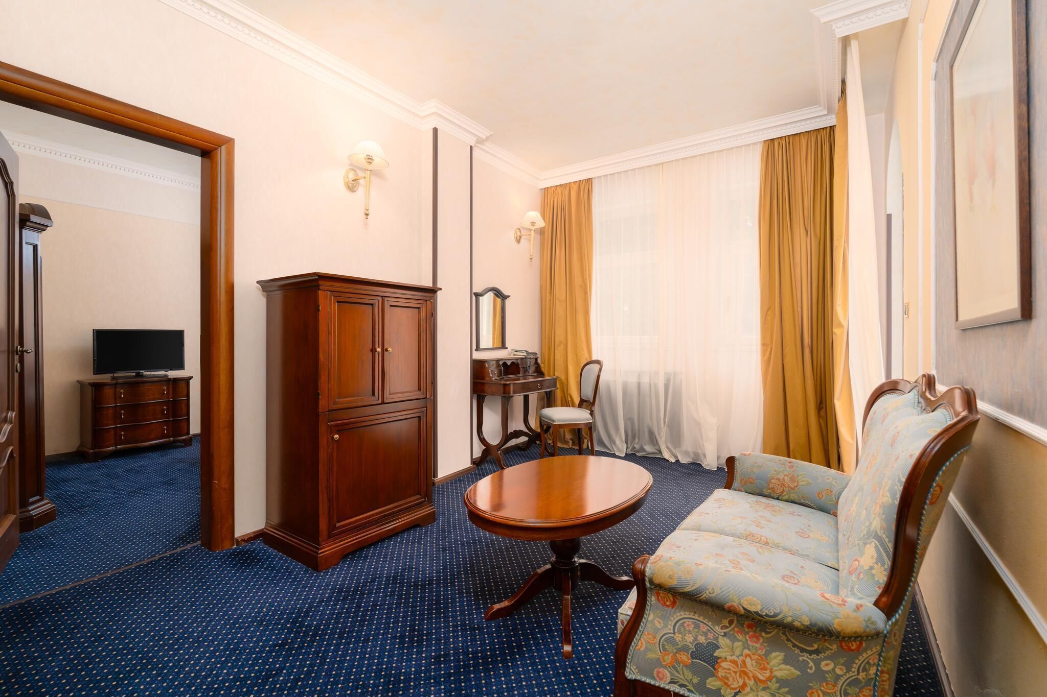 A hotel room with a blue carpet, a wooden cabinet, a round wooden table, a floral sofa, a small wooden desk with a chair, and a large mirror on the wall.
