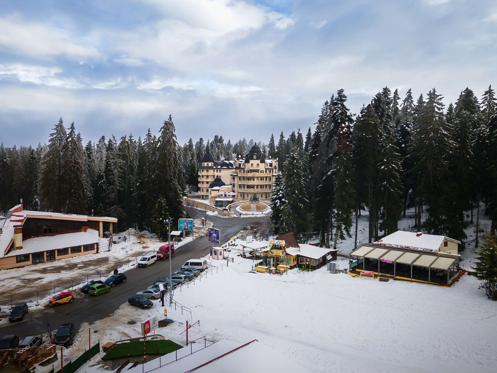 A snow-covered parking lot with several cars, a building with multiple floors, and a large tree in the foreground.