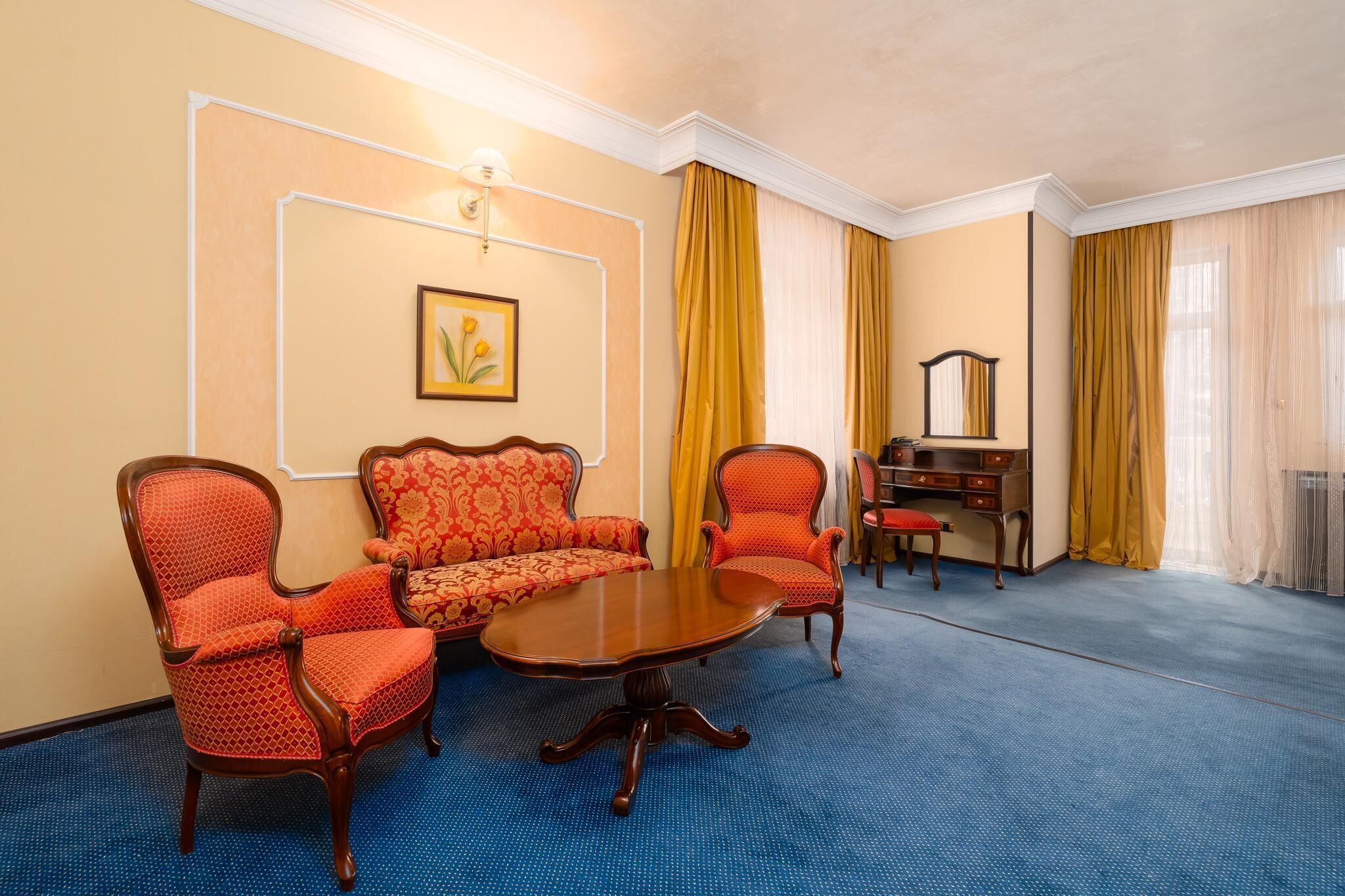 A hotel room with a blue carpet, a wooden coffee table, two red patterned chairs, a sofa, a wooden dresser, and a mirror.