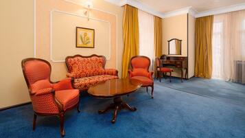A hotel room with a blue carpet, a wooden coffee table, two red patterned chairs, a sofa, a wooden dresser, and a mirror.