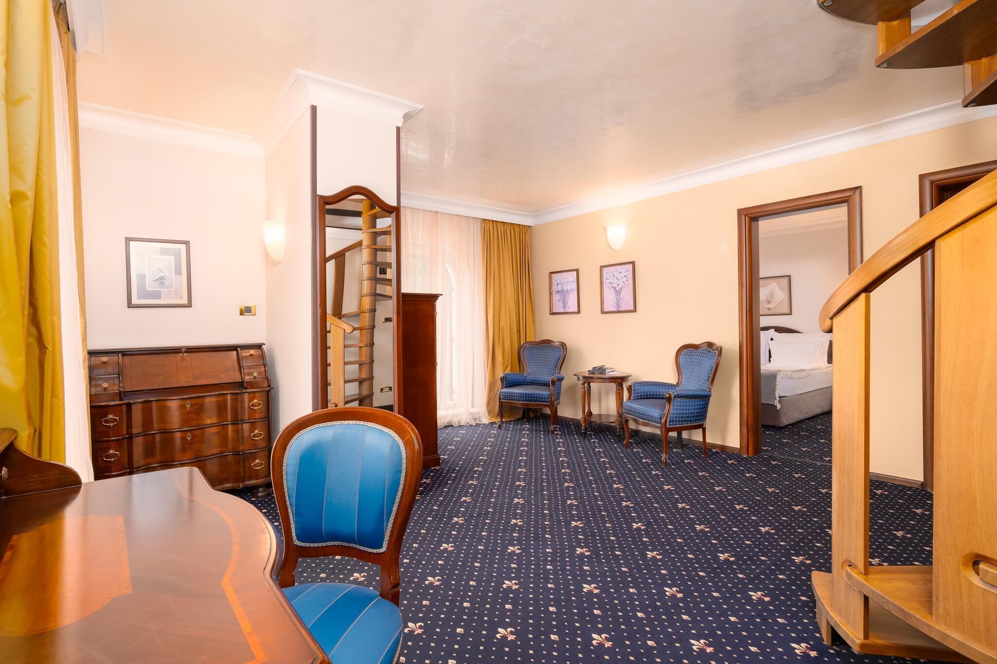 Une chambre d’hôtel avec un bureau en bois, deux fauteuils bleus, une petite table ronde, un lit et un escalier.