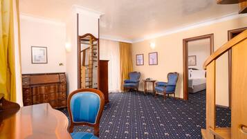 Habitación de hotel con un escritorio de madera, dos sillas tapizadas en azul, una mesita redonda pequeña, una cama y una escalera.