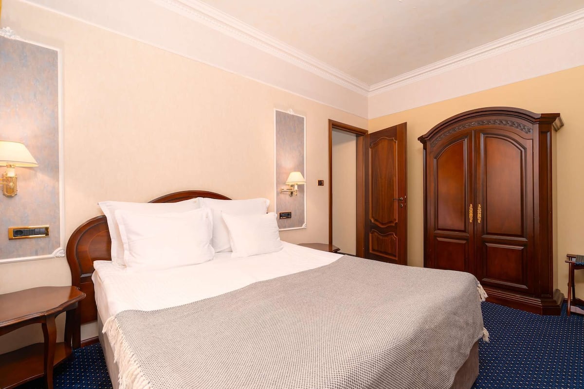 A hotel room with a large bed, a wooden wardrobe, a desk, and two wall-mounted lamps.