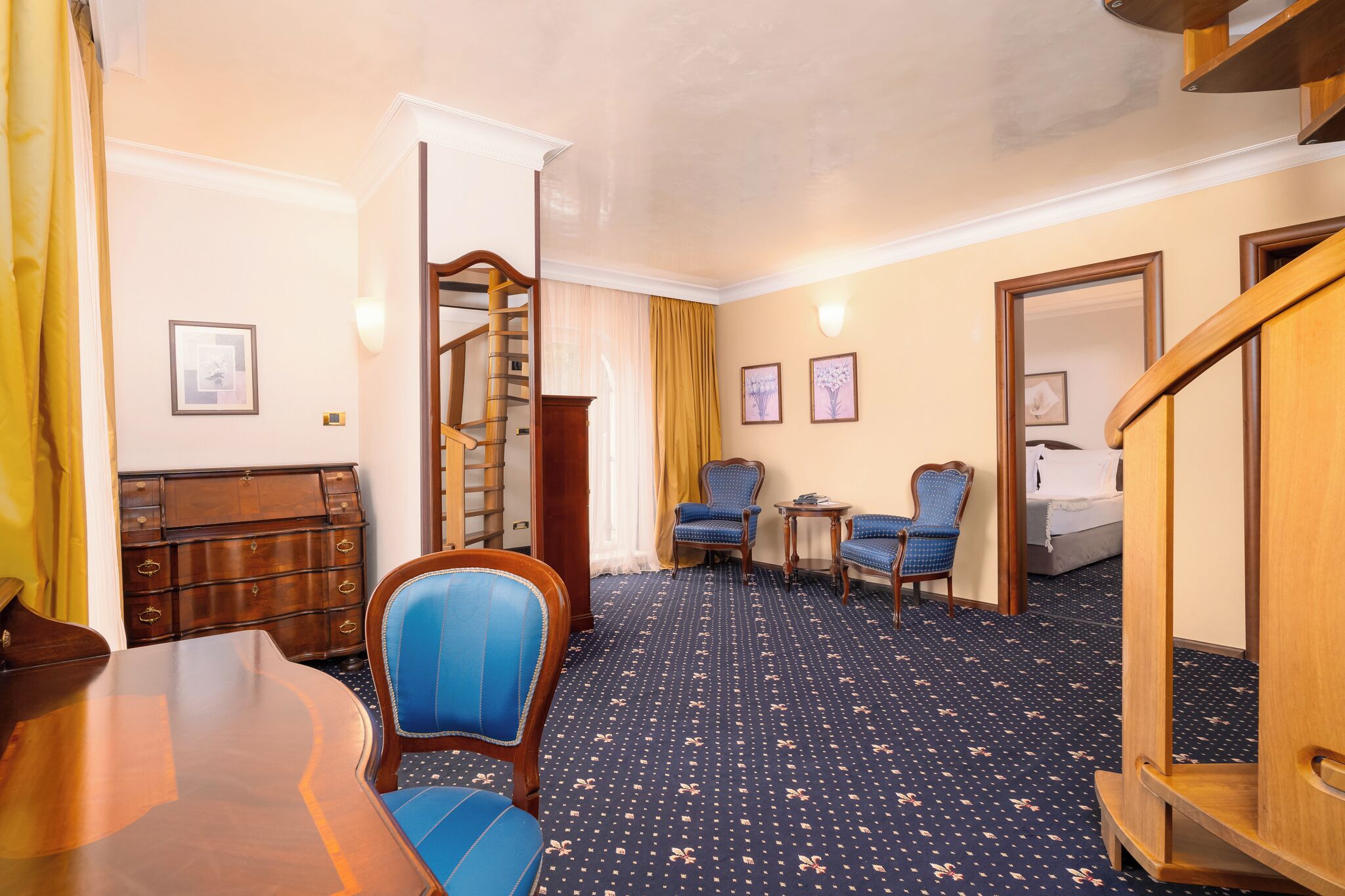 Ein Hotelzimmer mit einem Holztisch, zwei blau gepolsterten Sesseln, einem kleinen runden Tisch, einem Bett und einer Treppe.