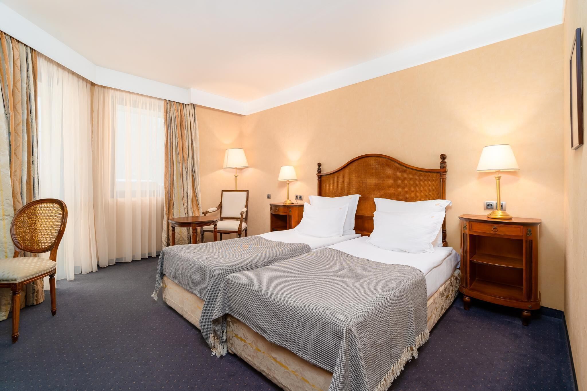 Quarto de hotel com uma cama de casal, duas mesas de cabeceira com luminárias, uma cadeira e uma janela grande com cortinas.