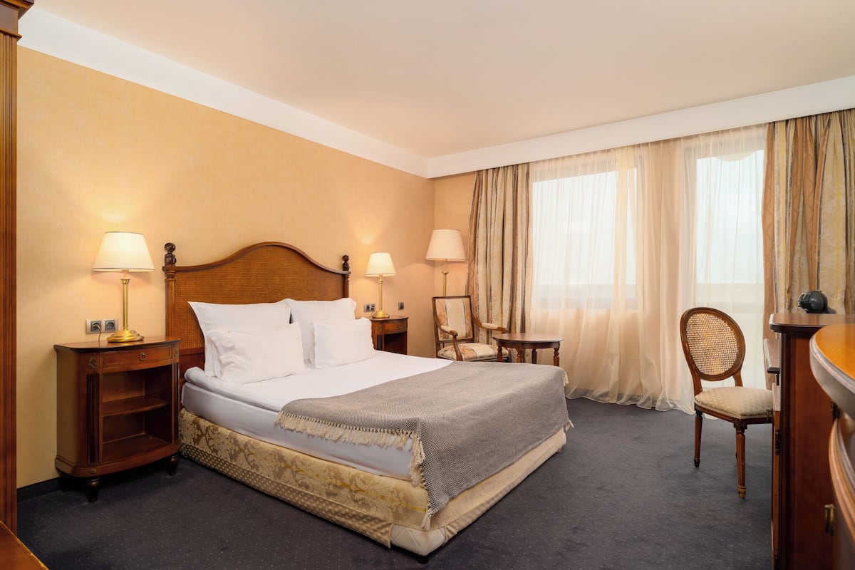 A hotel room with a large bed, two bedside tables with lamps, a chair, a small table, and a wardrobe.