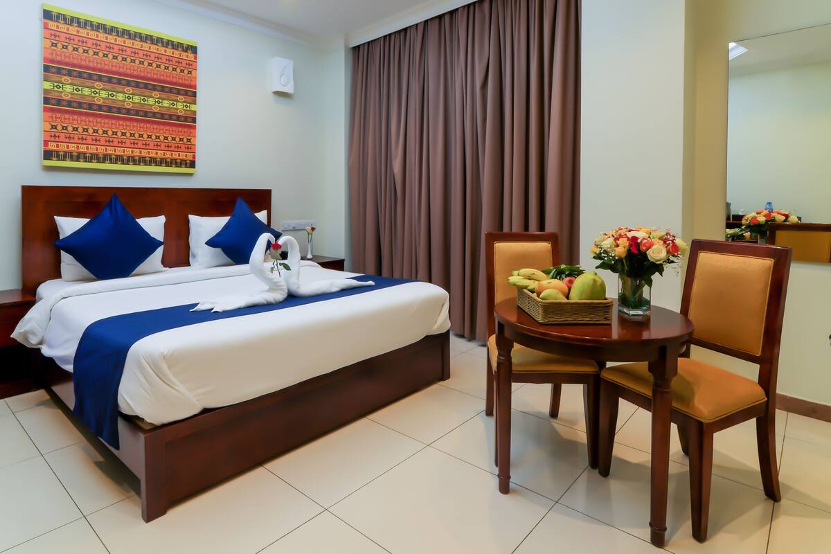 A hotel room with a neatly made bed, a dining table with fruit and flowers, and a large window with curtains.