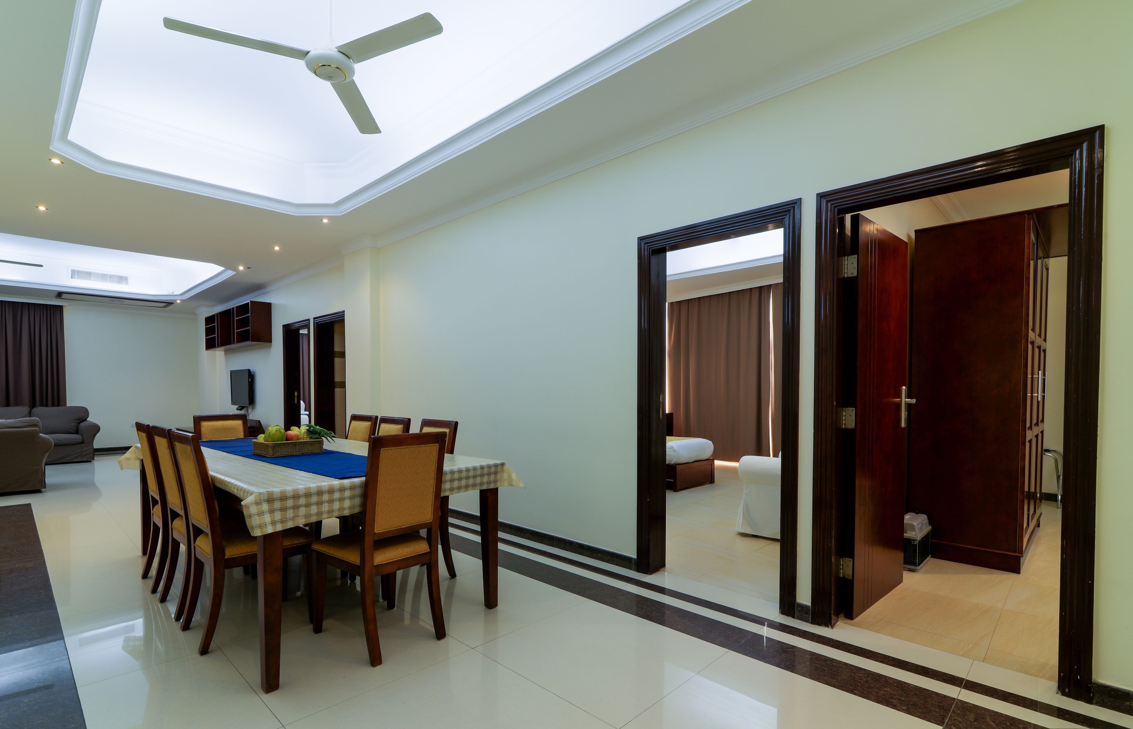 A spacious dining area with a high ceiling and skylight, a modern kitchen in the background, and a view into a bedroom through an open doorway.