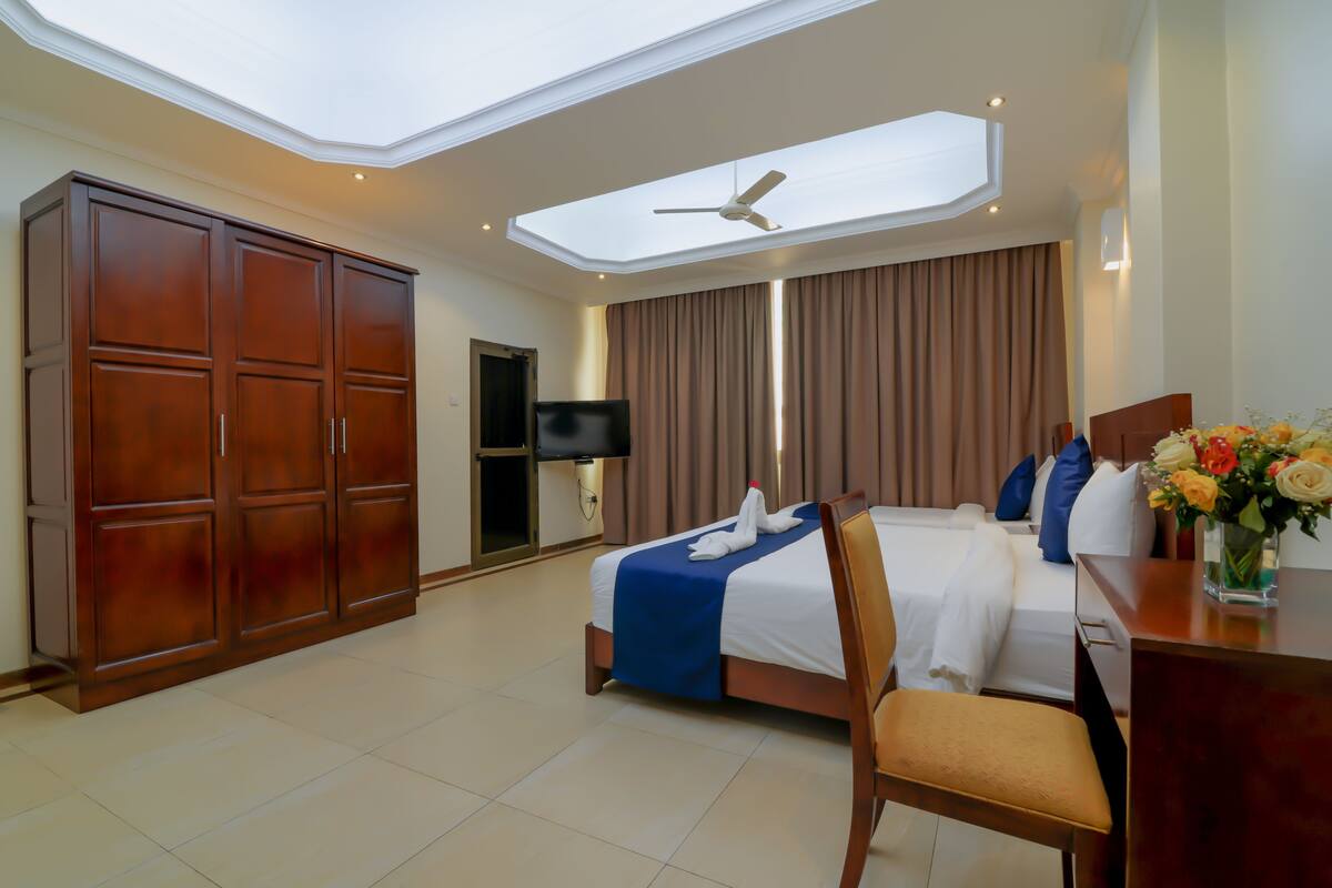 A hotel room with a skylight, a bed with a blue bedspread, a wooden wardrobe, a desk with a vase of flowers, and a television.