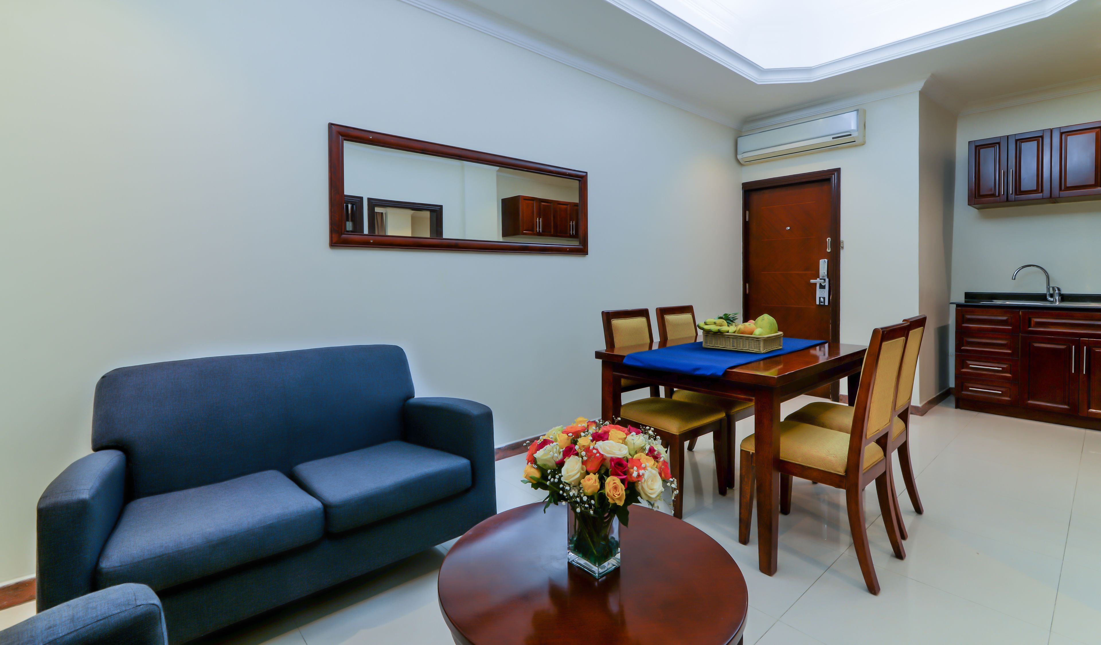 A living room with a blue sofa, a dining table with chairs, a vase of flowers, and a kitchen area with wooden cabinets and an air conditioning unit.