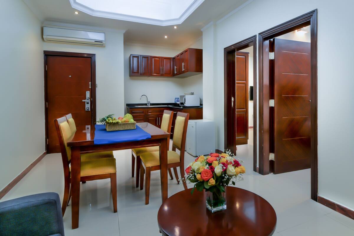 A dining area with a table, chairs, and a vase of flowers. There is a kitchen area with cabinets and a microwave in the background.