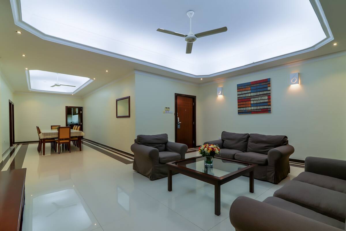 A spacious living area with a high ceiling, ceiling fans, a dining area with a table and chairs, and a sofa set.