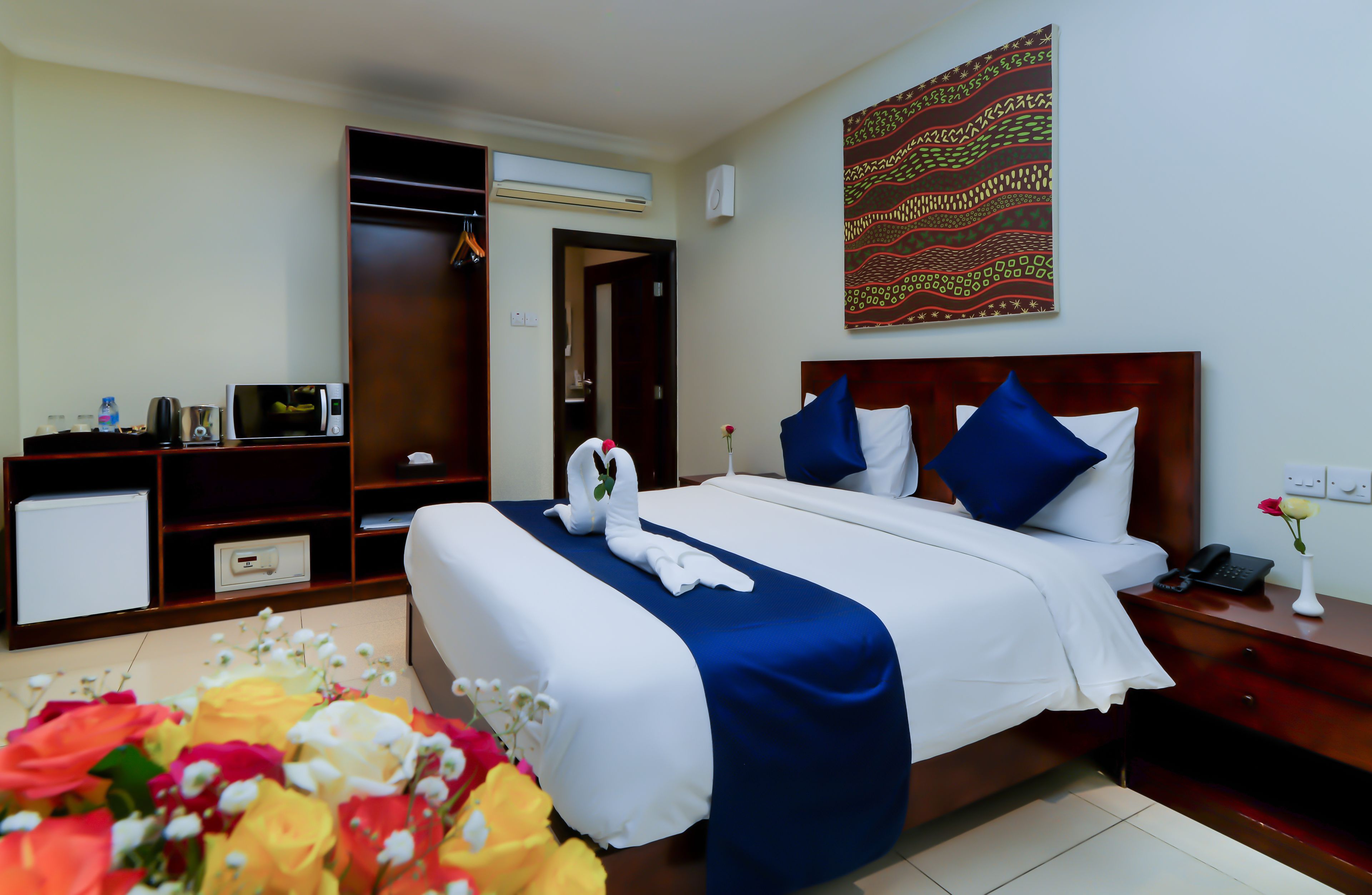 A hotel room with a neatly made bed, a bedside table with a phone and a vase of flowers, a small desk with a microwave, and a wardrobe.