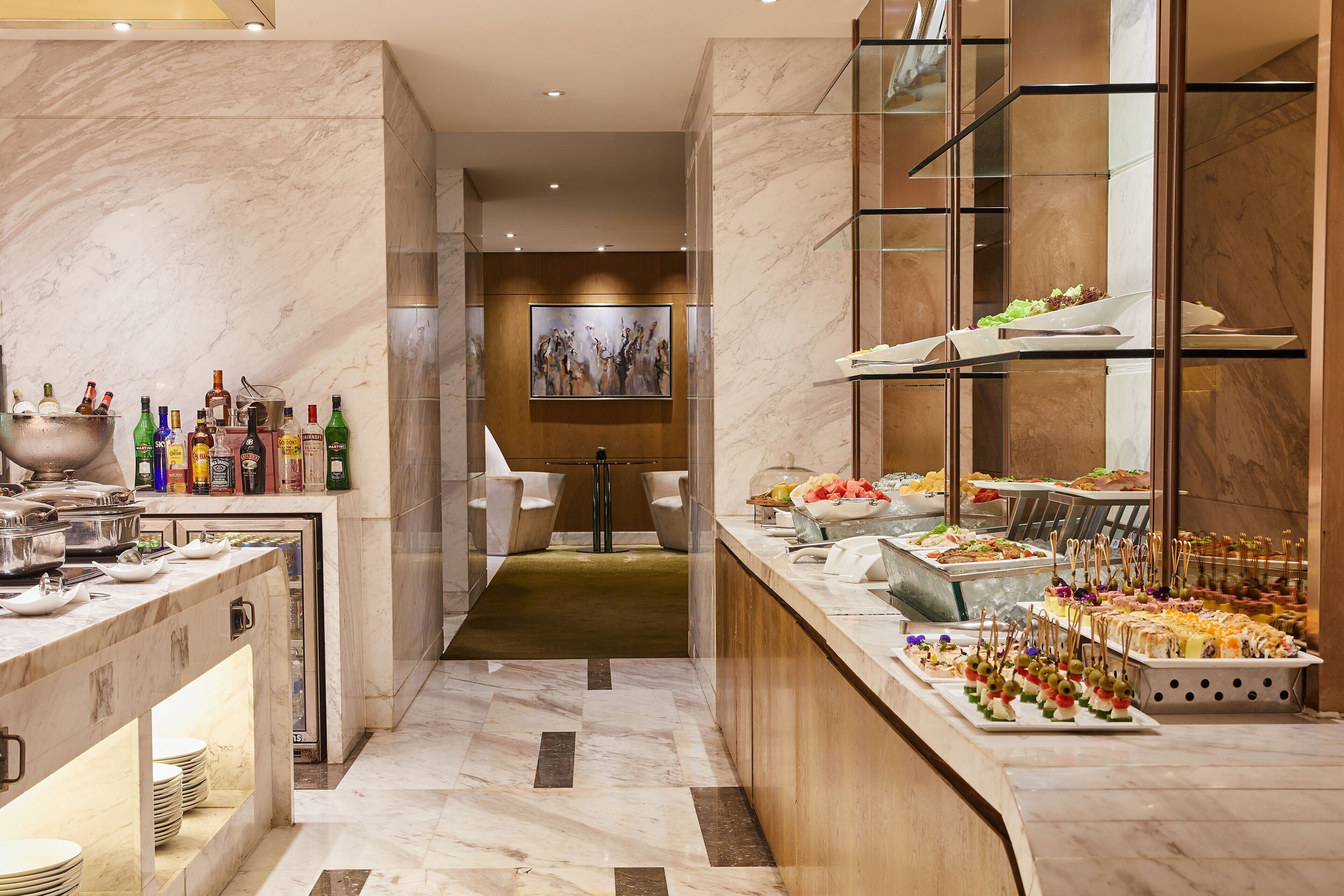 Un hall de hotel con un mostrador de recepción de mármol, una zona de buffet con diversas opciones de comida y un área de descanso con sillas blancas.