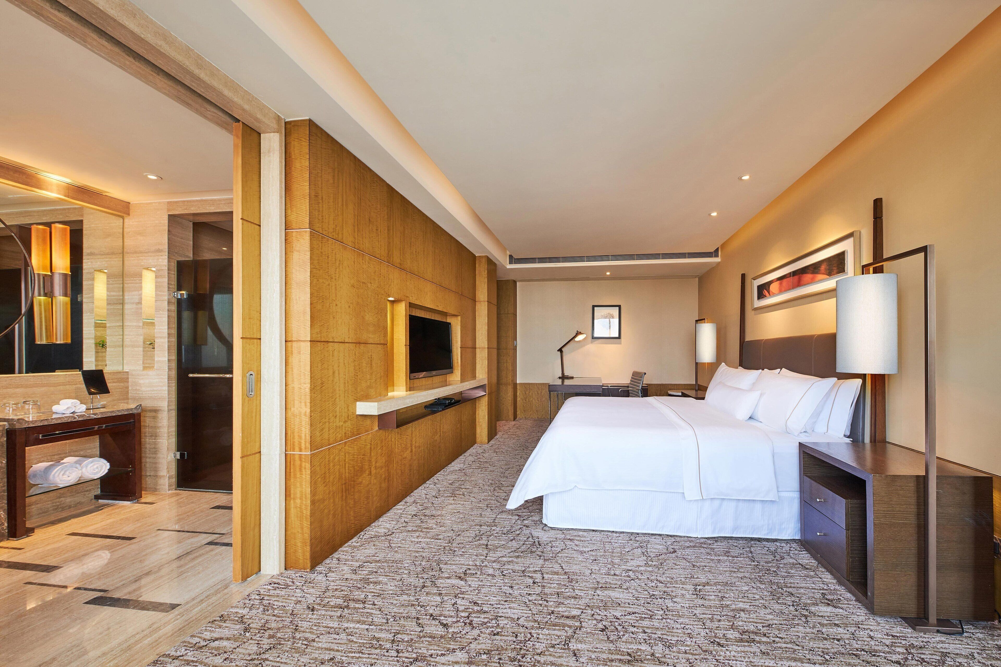 A hotel room with a large bed, a desk, a chair, a TV, and a bathroom with a mirror and towel rack.