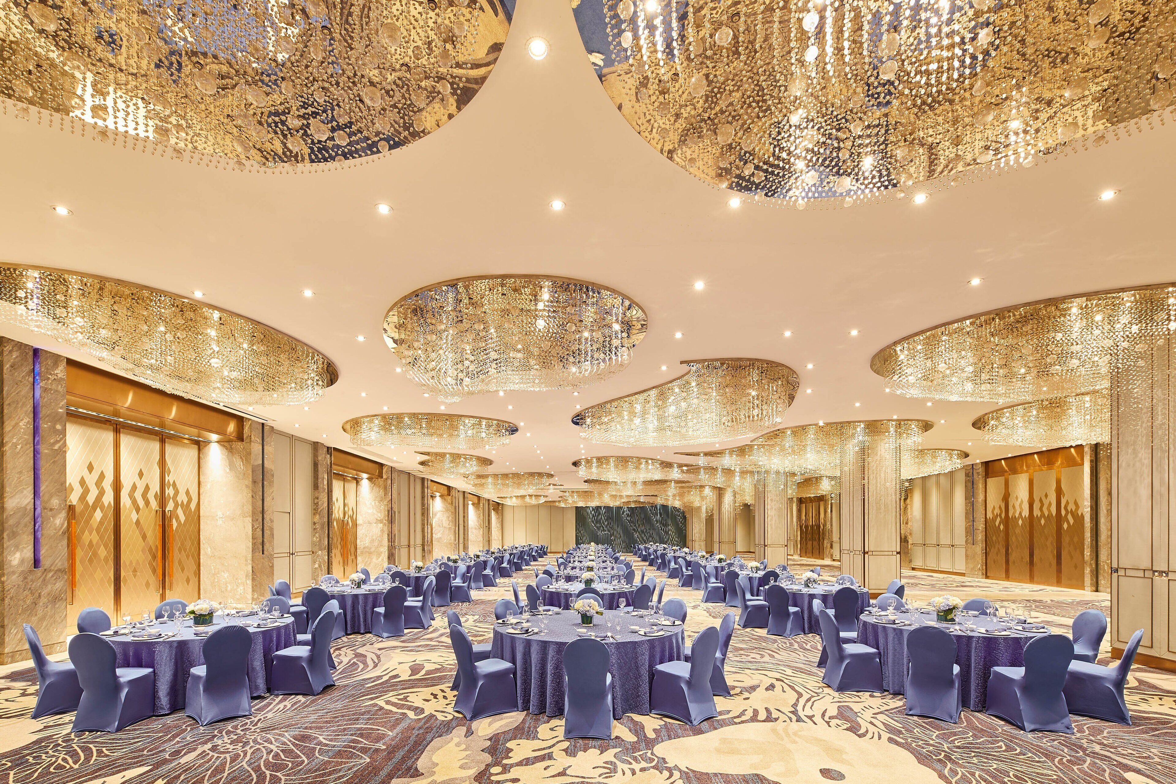 A large banquet hall with round tables, blue chairs, and ornate chandeliers.