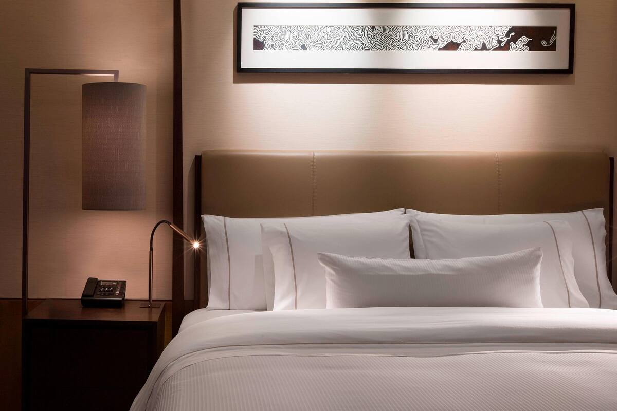 Un lit bien fait, avec du linge de lit blanc et une tête de lit, une lampe de chevet et une œuvre d’art encadrée accrochée au mur.