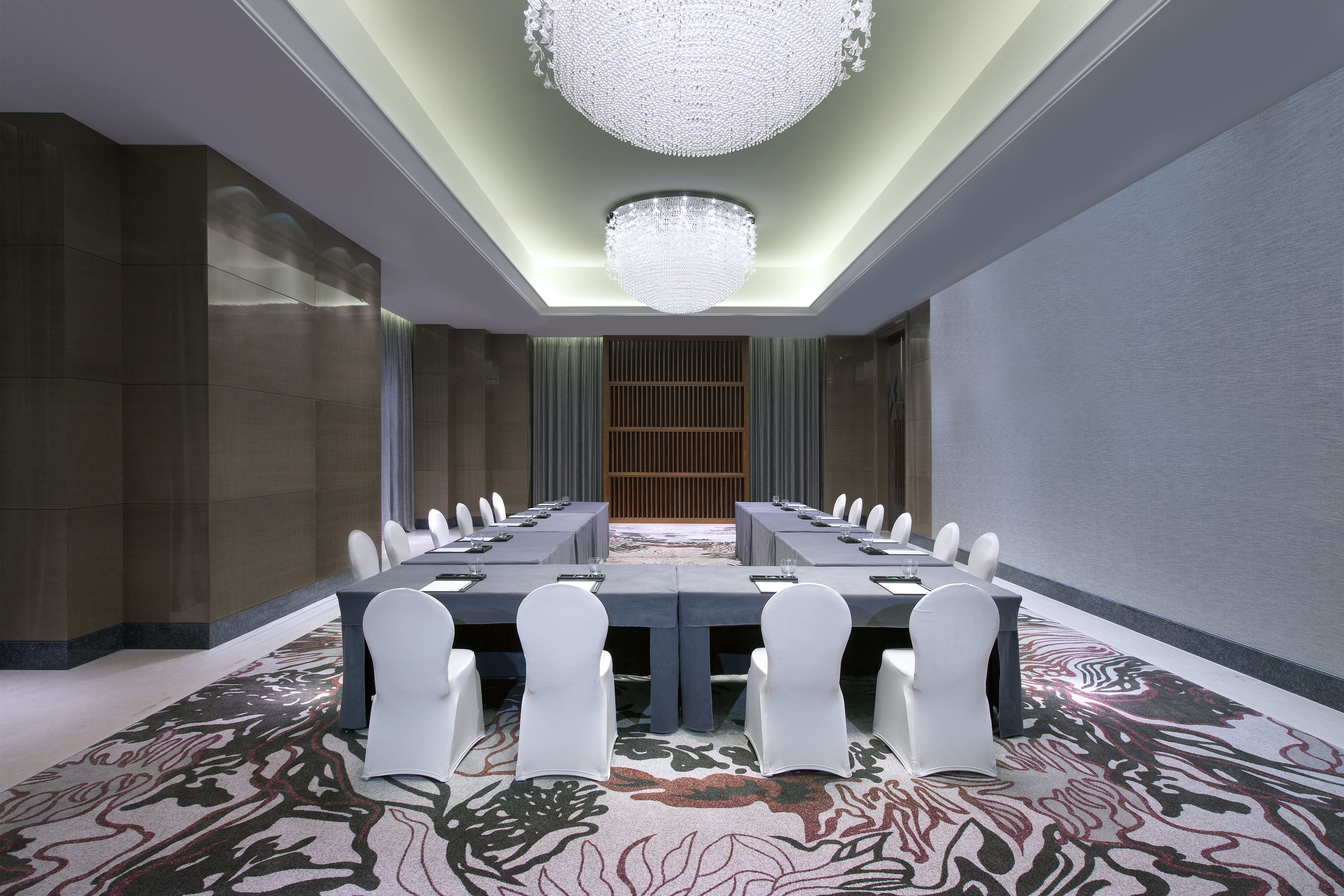 A conference room with a long table, white chairs, a chandelier, and patterned carpet.