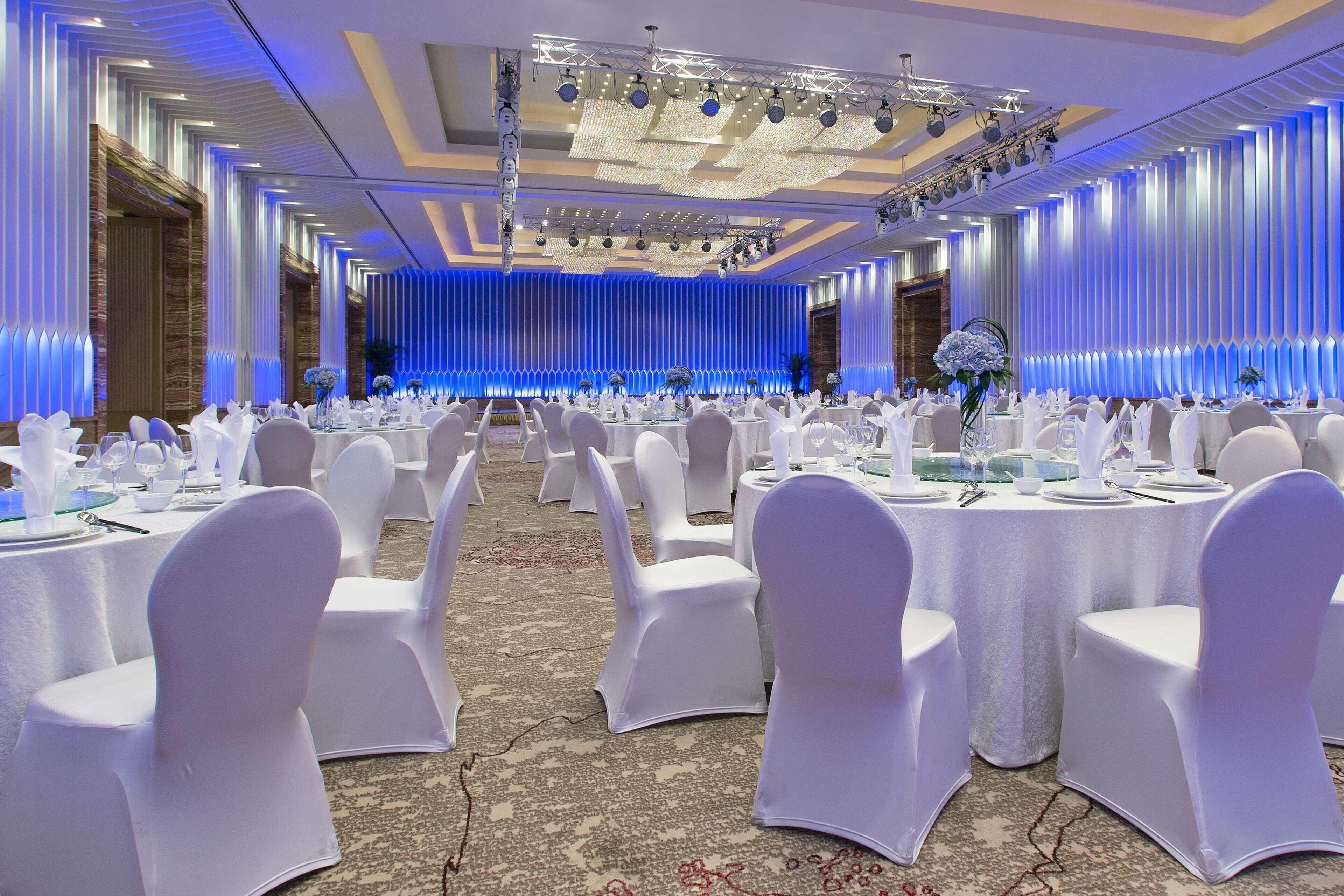 Un salón de banquetes con mesas redondas, sillas blancas y un escenario con iluminación azul.