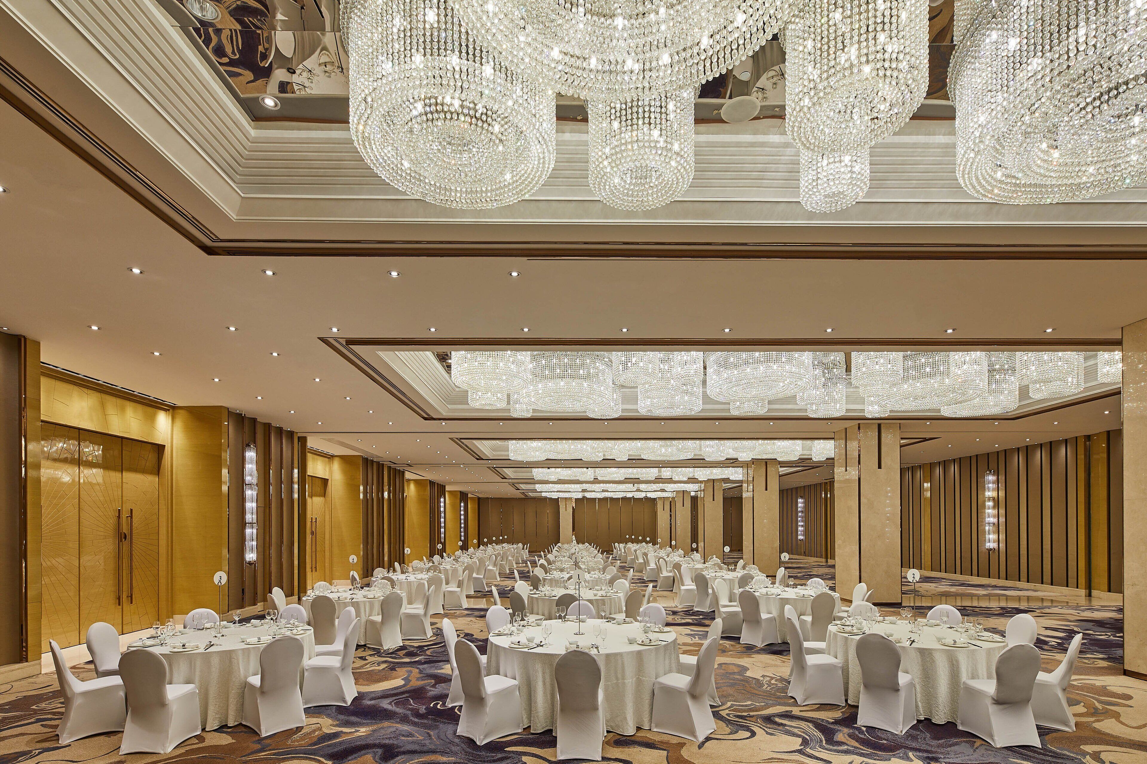 A large banquet hall with round tables, white chairs, and chandeliers.