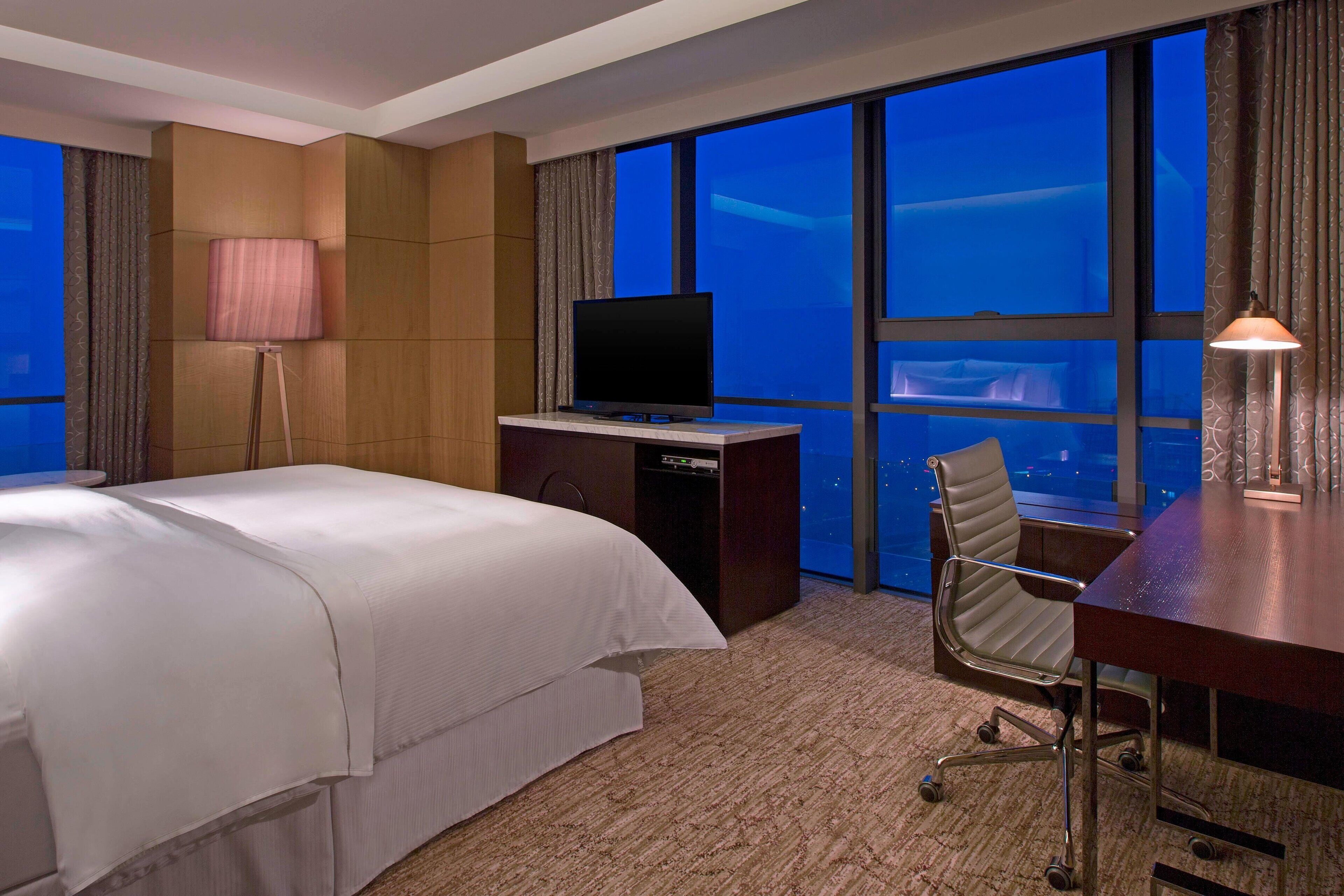 Habitación de hotel con cama, escritorio con televisor, silla y ventanas amplias con vista a la ciudad.