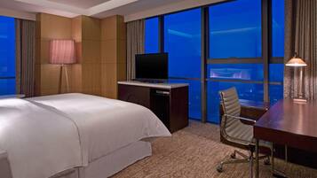 Habitación de hotel con cama, escritorio con televisor, silla y ventanas amplias con vista a la ciudad.